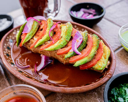 Delicious Guadalajara Mexican style torta ahogada of carnitas, accompanied by red onion, salsa, avocado and tomato, served in a clay dish.の写真素材