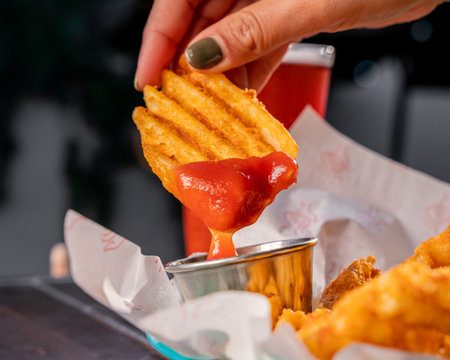 Hand holding a hot, crunchy crisscut and spreading ketchup on a craft beer counter.