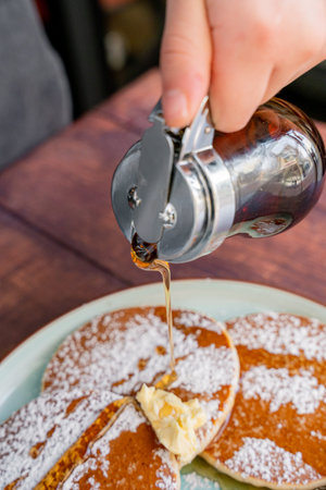 person putting maple syrup on their delicious and fluffy buttery pancakes, accompanied by green...