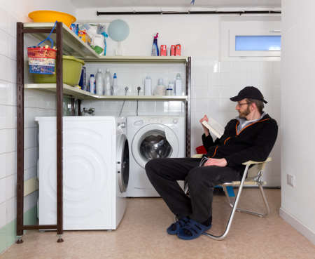 Man reading a book while waiting for the laundry to wash in a home laundry roomの写真素材