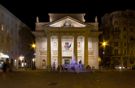 TRIESTE, Italy - August 3, 2017: Night view of the Palazzo della Borsa Vecchia in Piazza della Borsa in Trieste, Italy, with people walking by and sitting on the fountain in front of the palaceのeditorial素材