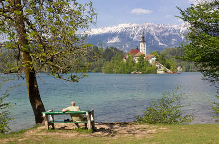 BLED, Slovenia - April 25, 2018: Man sitting on a bench on the shoreline contemplating the church on the Bled islandのeditorial素材