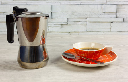 Orange colored cup of coffee with a modern design and a coffee maker on a white wooden tableの写真素材