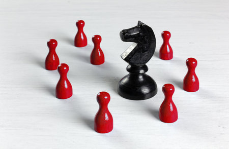 Big black knight chess piece surrounded by smaller red pawns on a white wooden table