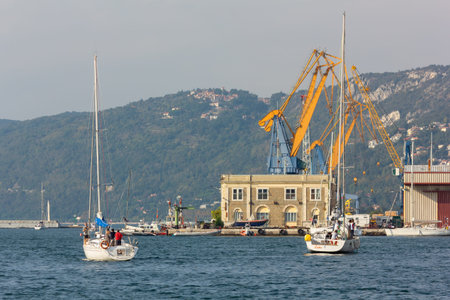 TRIESTE, Italy - October 12, 2014: Two sailboats in the waters of the gulf, in front of a few seaport's buildings and cranesのeditorial素材