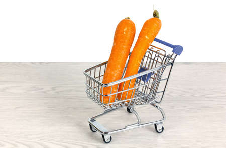 Two carrots inside a miniature shopping cart on a white wooden table over a white backgroundの写真素材