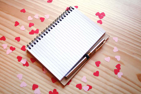 St Valentine's day: a notepad to write a love note for the loved one, with a pen, pink and red hearts on a wooden table. Top view with copy spaceの写真素材
