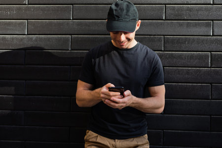A man on a wall background in a black t-shirt and baseball cap looks into a smartphone