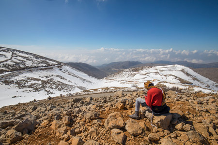 A young man sitting on a hill overlooking snowy mountains
