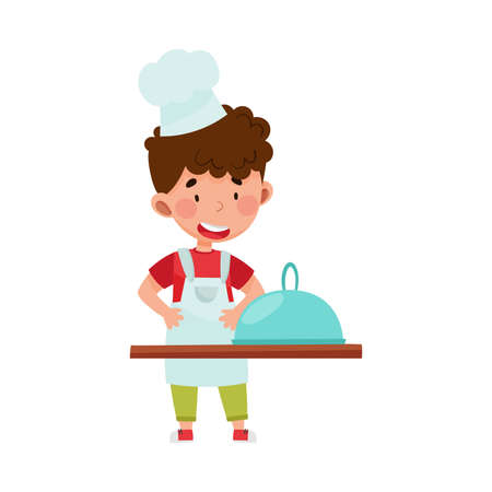 Little Boy Character in Hat and Apron Standing at Kitchen Table Cooking Dish Vector Illustration. Smiling Kid Chef Engaged in Cookery Conceptのイラスト素材