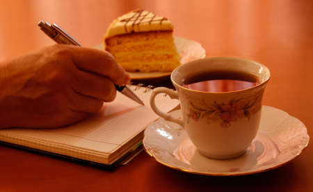 Hand writing on notebook, cup of tea and piece of cake on wooden backgroundの写真素材