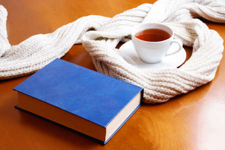 White cup of tea, gray scarf and closed blue book on wooden tableの写真素材