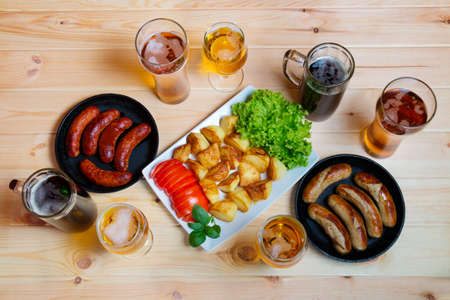 Mugs of beer and glasses of beer, plate of roasted potato and frying pans with grilled sausagesの写真素材