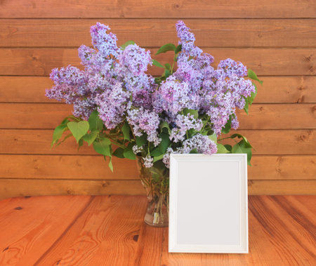 Lilac flower in glass vase and white frame on wooden background