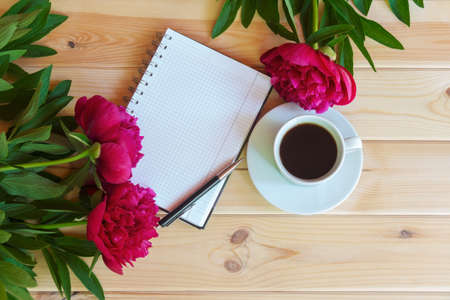 Morning coffee cup for breakfast, empty notebook and red peony flowers on wooden table. Top view, copy space.の写真素材