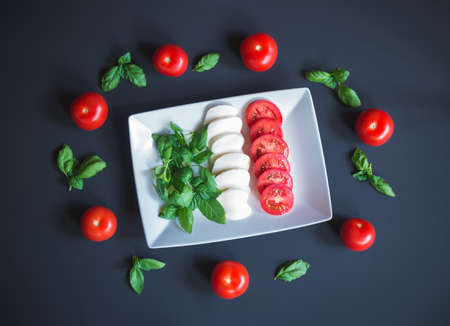 Mozzarella, tomato, basil in form and color of italian flag on white plate with tomatoes and basil leafes aroundの写真素材