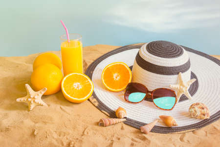 Glass of orange juice, hat, sunglasses and seashells on sand beach on blue backdropの写真素材
