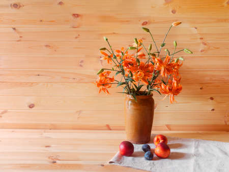 Bouquet of orange lilies in clay vase and fresh fruits near. Wooden background. View with copy space.の写真素材