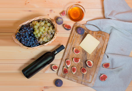 Red wine bottle, figs, piece of cheese, bunches of grape in wicker basket and honey in glass bowl ...