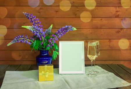 Blue wildflowers lupine, glasses of champagne, empty white frame and  golden gift box on wooden background. Selective focus. View with copy space.の写真素材