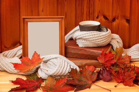 Mug of coffee wrapped in gray woolen scarf, empty wooden frame, autumn leaves and stack of books on wooden table. View with copy spaceの写真素材