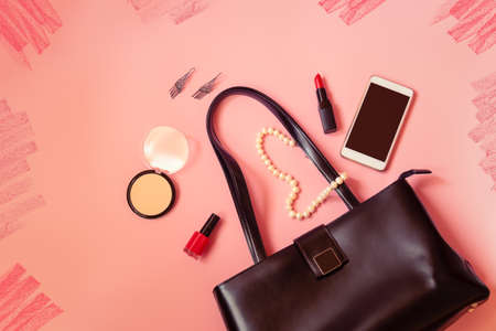 Black leather woman's bag, smartphone, earrings, face powder, lipstick and nail polish on pink background. Top view with copy space.の写真素材