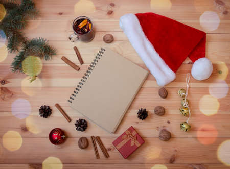 Empty notebook, fir branch, flutty Santa hat, christmas decorations, glass of mulled wine and red gift box on wooden background.  Top view, copy space.の写真素材