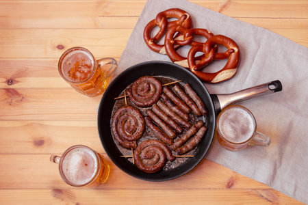 Frying pan with roasted sausages, beer mugs and soft salted pretzels on wooden table. Top view.