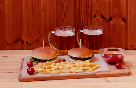 Two mugs of beer, burgers and fried potatoes on wooden table. Selective focus.