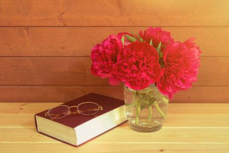 Red peonies in vase, book and glasses on wooden table. View with copy space.