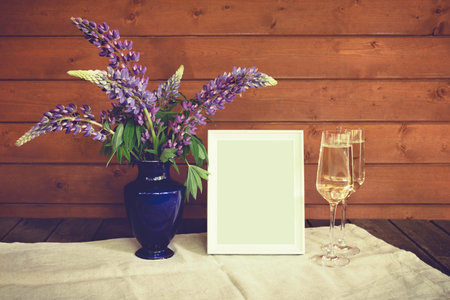 Wildflowers lupine, glasses of champagne and empty white frame  on wooden table. Toned image. View with copy space.の写真素材