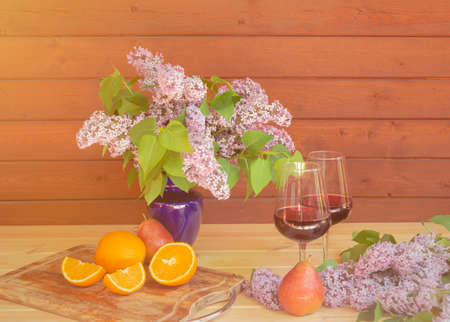 Flowers of lilac in blue vase, glasses of wine and fresh fruits on wooden table.の写真素材