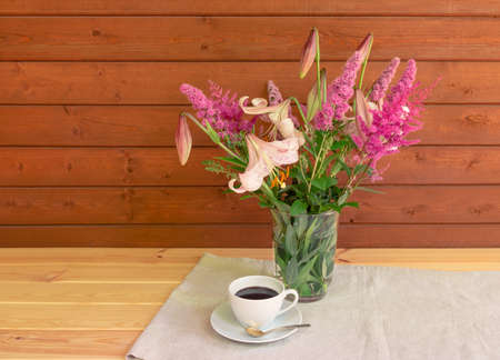 Bouquet of beige colored lilies with pink spots, pink spirea and astilbe flowers in glass  vase and cup of coffee on wooden table.の写真素材