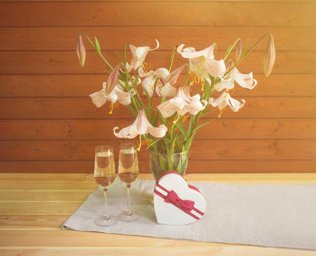 Bouquet of beige colored lilies with pink spots in glass  vase, two glasses of champagne and heart shape gift box on wooden table.の写真素材