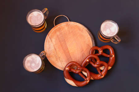 Beer mugs and soft salted pretzels around cutting board on dark background. Top view, copy space.の写真素材
