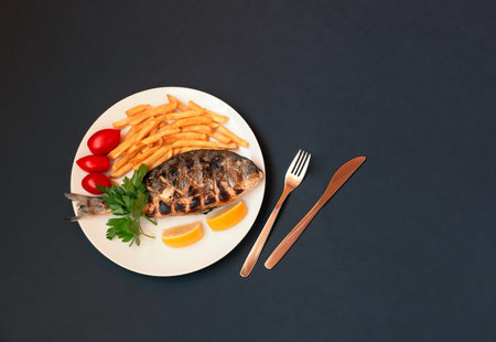 Delicious roasted dorado fish with lemon, fried potatoes, fresh parsley and tomatoes on white plate on dark background. View from above, copy space.の写真素材