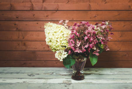 Autumnal fading pink and white hydrangea flowers in glass vase on aged wooden table. Toned image. View with copy space.の写真素材