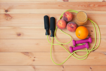 Pink dumbbells, jumping rope, fresh fruits and homemade healthy fruits smoothie made of oranges, peaches and bananas on wooden background. Healthy eating and fitness concept. Top view, copy space.の写真素材