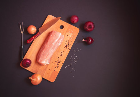Fresh pork fillet on cutting board on dark background. Knife, fork for meat and fresh onions near. Top view, copy space.の写真素材