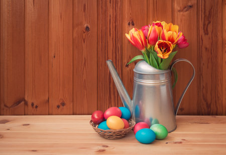 Easter concept. Colorful tulips in watering can and Easter eggs in wicker basket on wooden table....