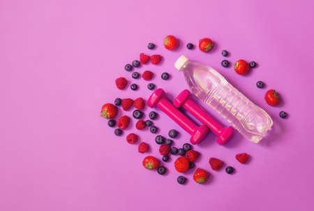 Pink dumbbells, bottle of water and berries on purple background. Healthy eating and fitness concept. Top view, copy space.の写真素材