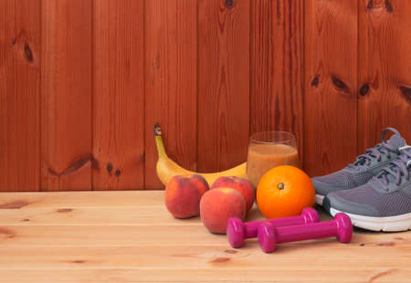 Fresh fruits, pink dumbbells, sneakers and homemade healthy fruits smoothie made of oranges, peaches and bananas on wooden background. Healthy eating and fitness concept. View with copy space.の写真素材