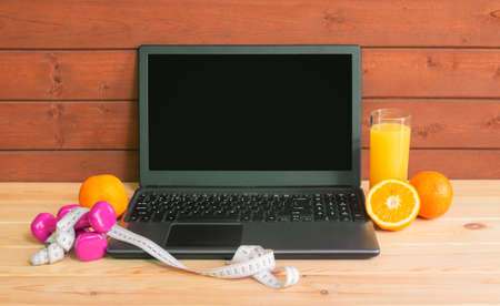 Laptop computer, dumbbells, measuring tape, glass of orange juice and fresh oranges. Exercises, healthy food, online fitness training, staying fit at home. View with copy space.の写真素材