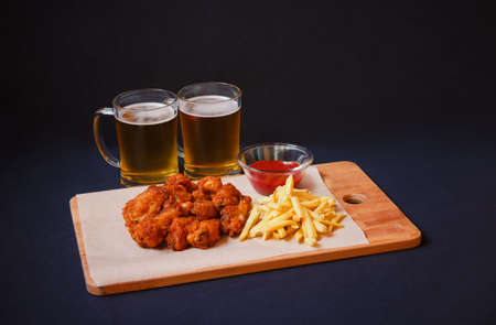 Chicken wings with french fries on cutting board and mugs of beer near on dark background. Selective focus. View with copy space.の写真素材