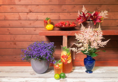 Cold summer lemonade with lemon, strawberry and lime, brown plate of strawberry, wicker basket with beautiful red  lilies and pink astilbe flowers on bench, pink astilbe flowers in blue vase and lobelia blue blossom in flower pot on aged wooden board.の写真素材