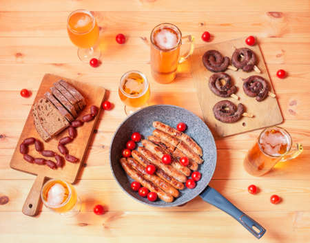 Frying pan with roasted sausages, roasted sausages on cutting board, beer mugs, beer glasses, bread and little chorizo sausages on wooden table. Top view.の写真素材