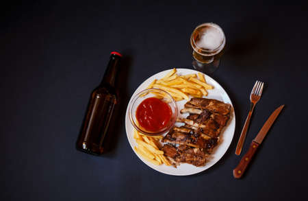 Baked pork ribs with french fries on white plate, bottle of beer and glass of beer near on dark background. Top view, copy space.の写真素材