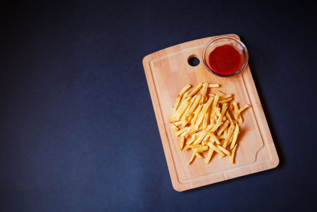 Tasty french fries and ketchup on cutting board on dark background. Top view, copy space.