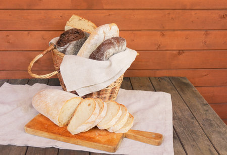 Assorted fresh bread in wicker basket and sliced wheat bread on cutting board near on aged wooden table.の写真素材