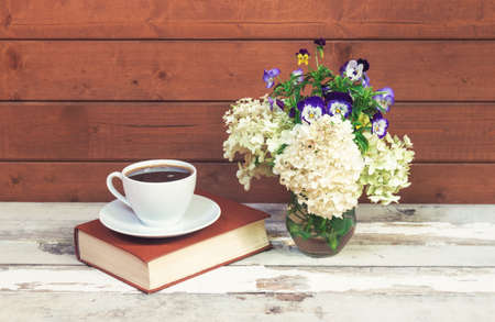 Cup of coffee, book and white hydrangea and pansy flowers in glass vase on aged wooden table. View with copy space.の写真素材